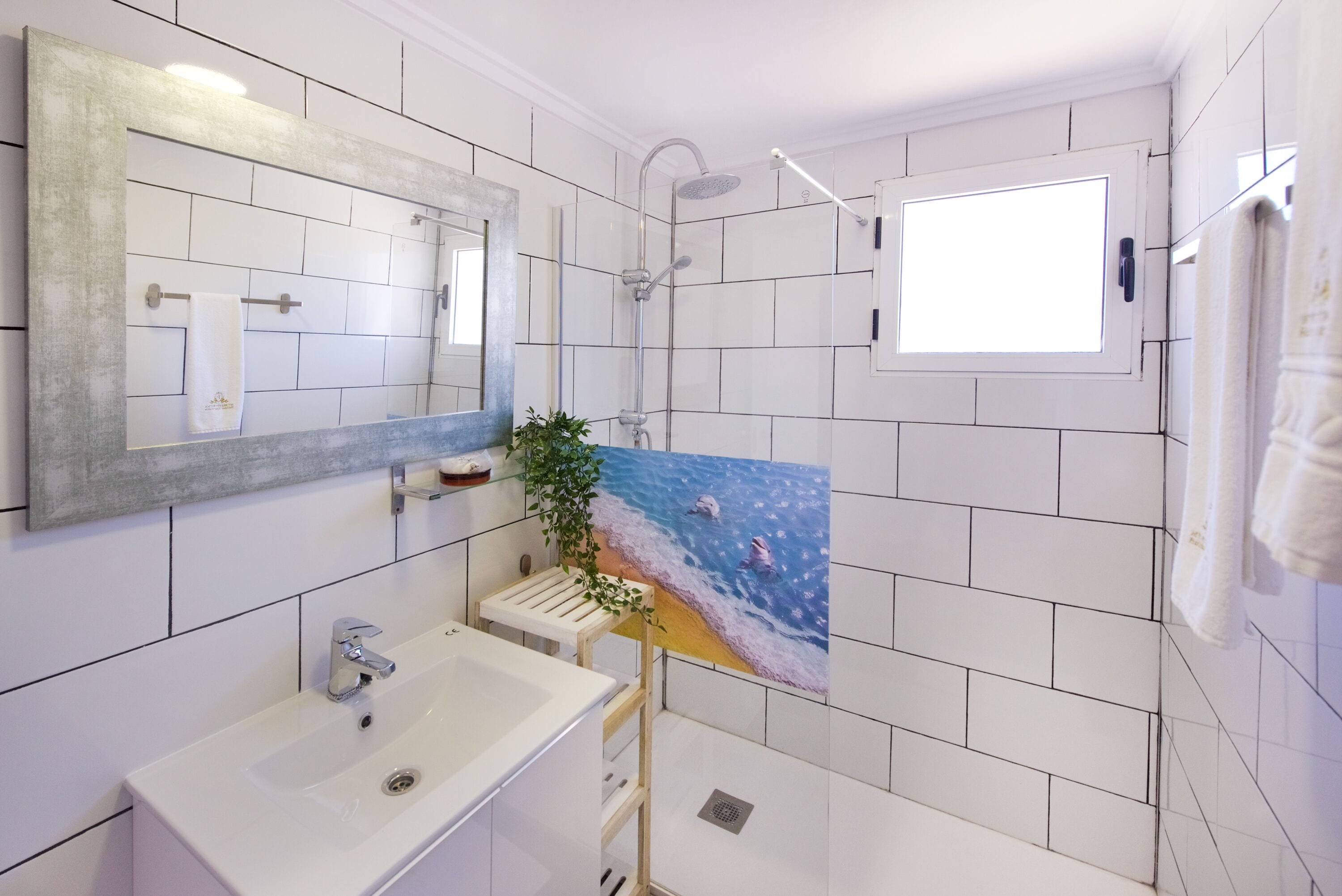 Een badkamer met witte tegels, een grote spiegel, een wastafel, een douche en een raam.