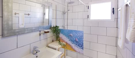 A bathroom with white tiles, a large mirror, a sink, a shower, and a window.