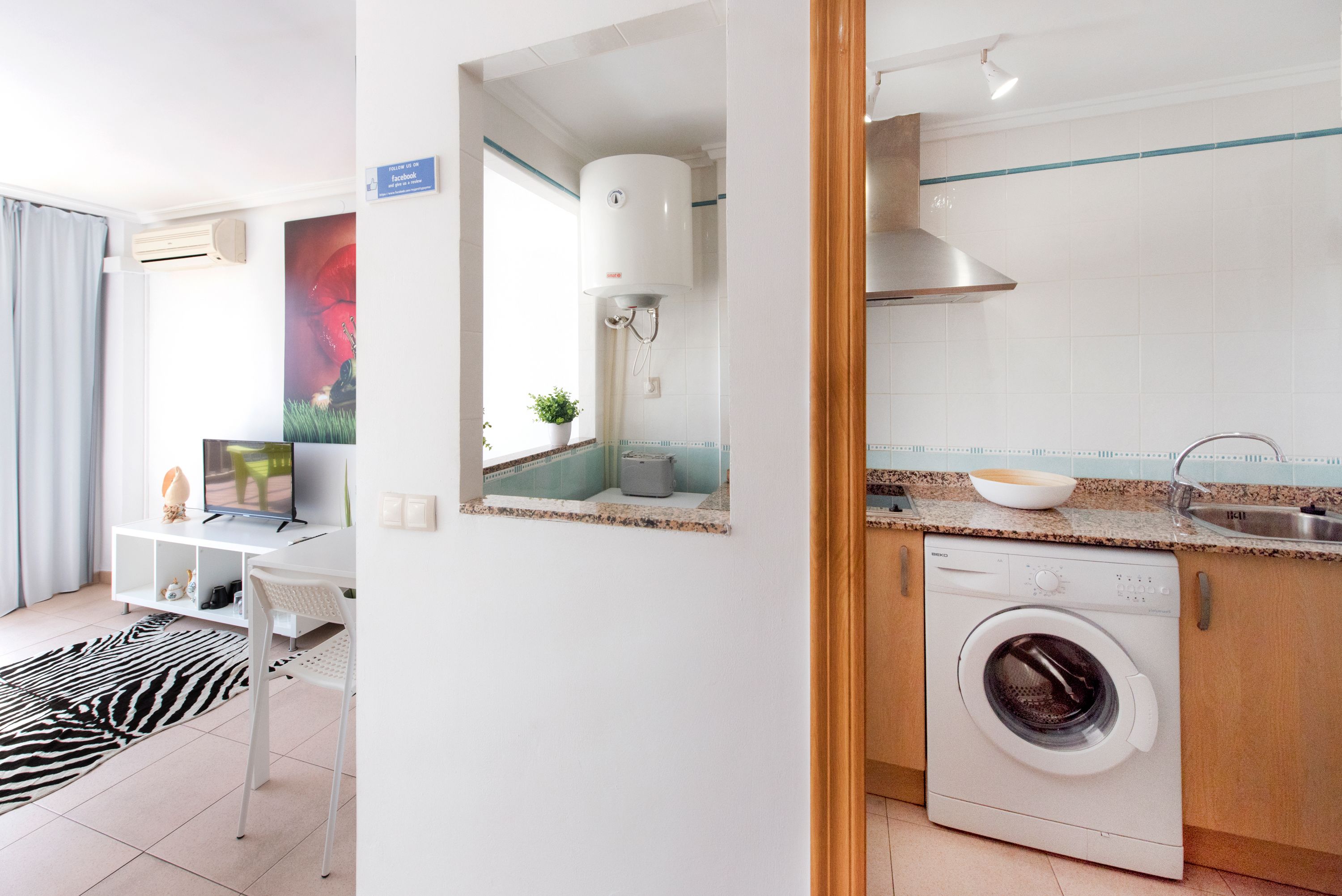 A modern kitchen with a built-in washing machine, a dining area with a white table and chairs, and a large mirror reflecting the kitchen.