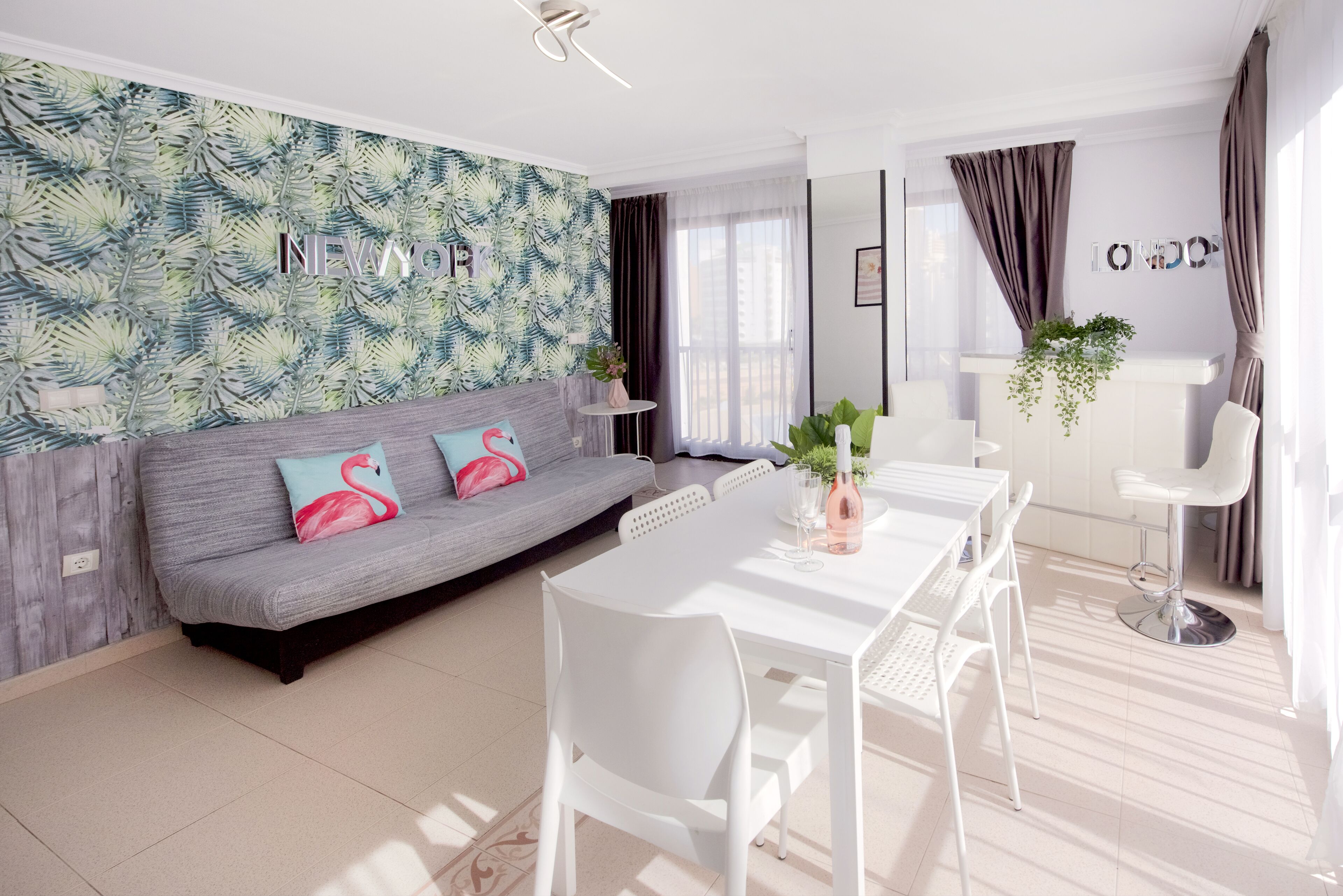 A modern living room with a grey sofa, white dining table, and chairs. There is a wall with a tropical leaf pattern and the word "NEW YORK" on it.