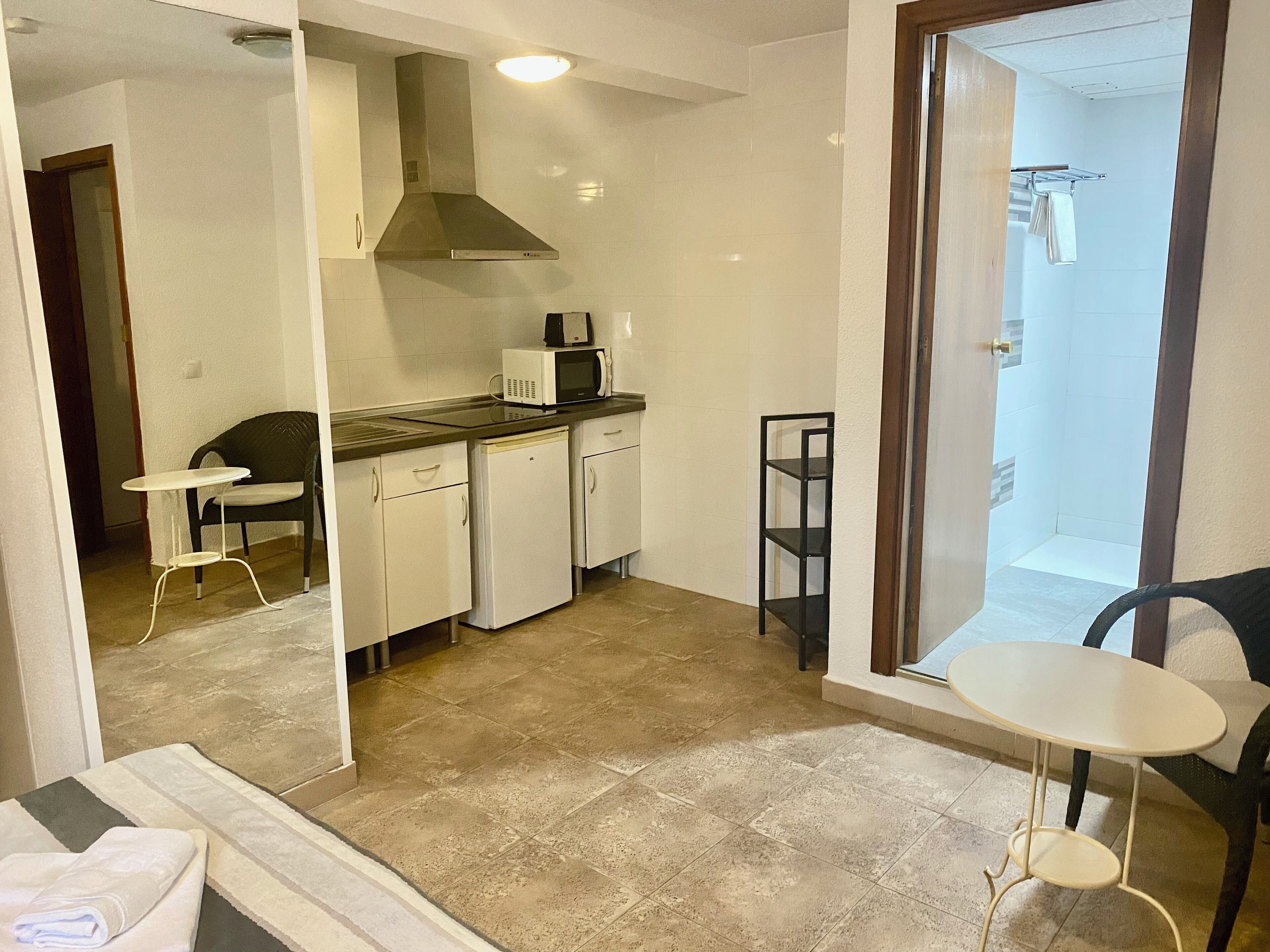 A compact kitchen with a microwave, a coffee maker, and a small refrigerator. There is a dining area with a table and chairs. The room has a tiled floor and a view of a shower area through a glass door.