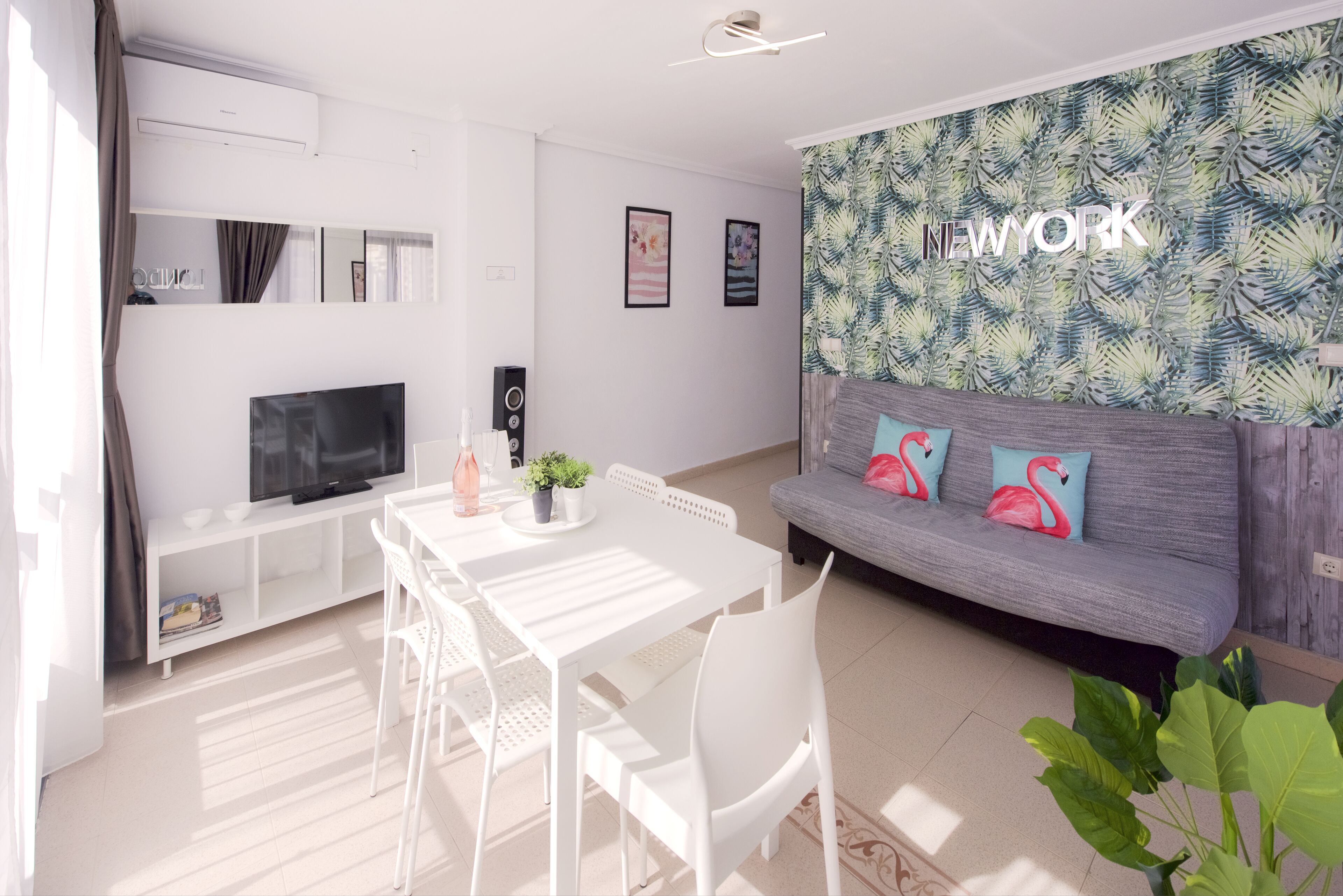 Una sala moderna con un sofá gris, una mesa de comedor blanca y sillas. Hay un televisor de pantalla plana sobre un mueble blanco, una planta pequeña sobre la mesa y una pared con un estampado de hojas tropicales.