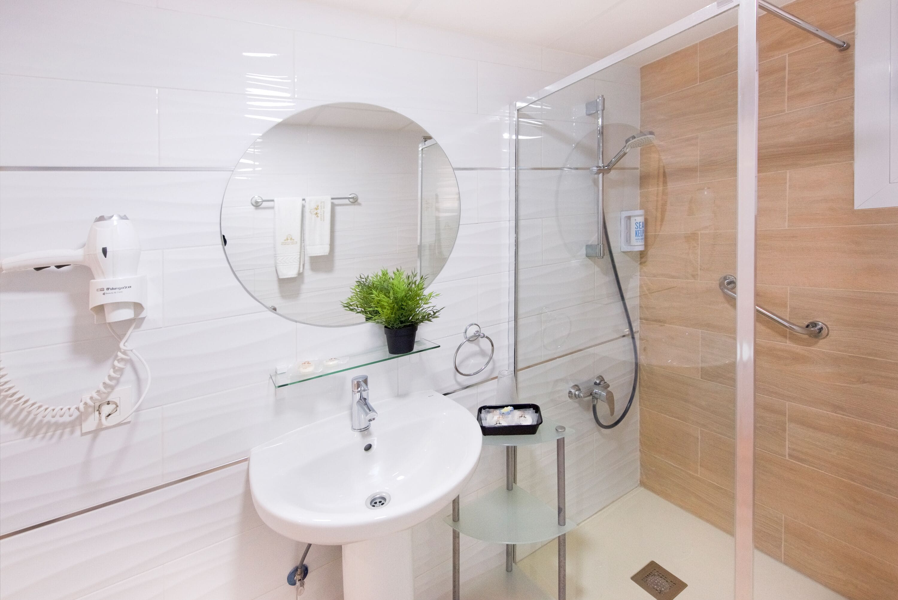 Un bany modern amb dutxa de vidre, un lavabo blanc i un mirall rodó.