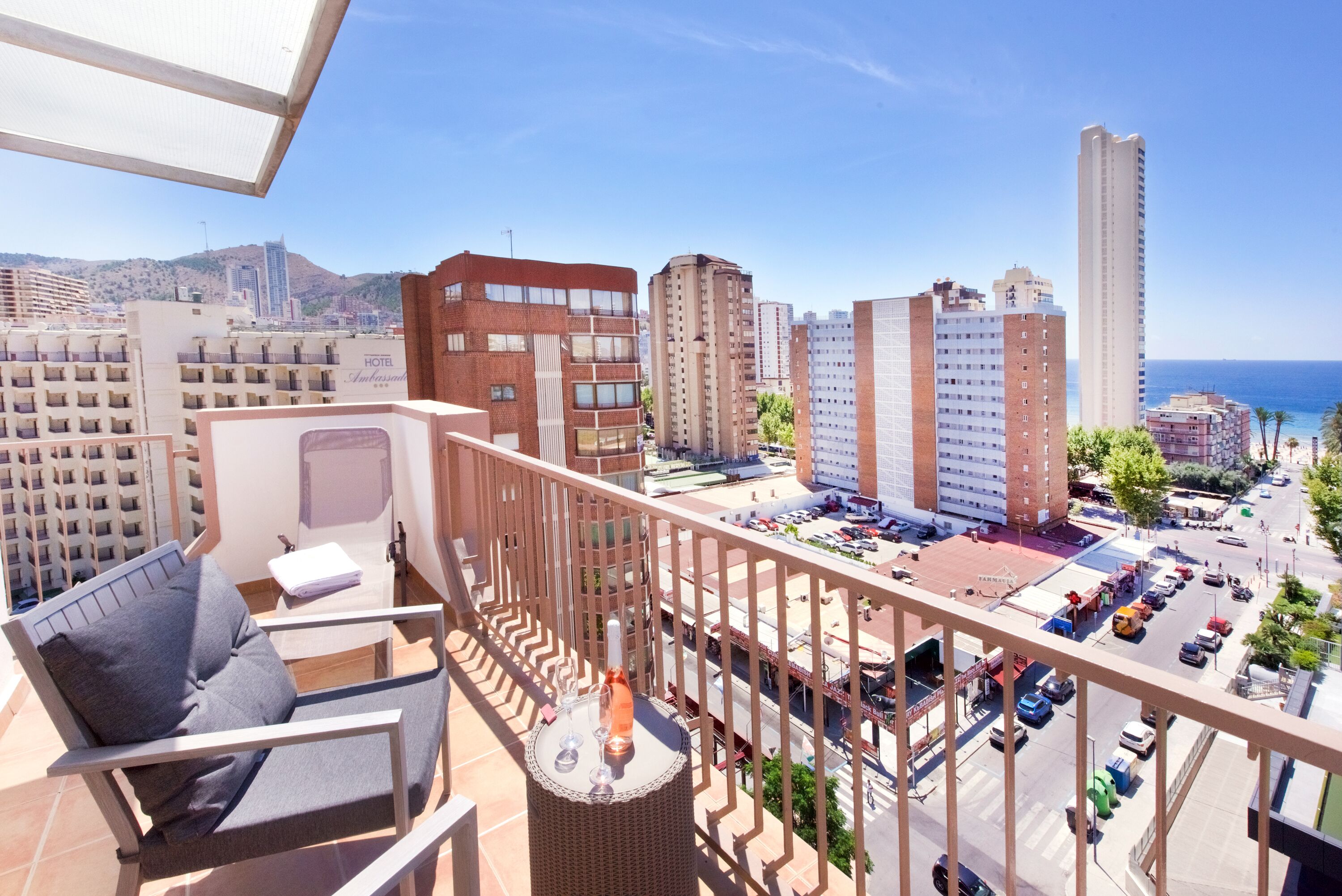 A balcony with a chair and table, overlooking a cityscape with buildings and a clear sky.