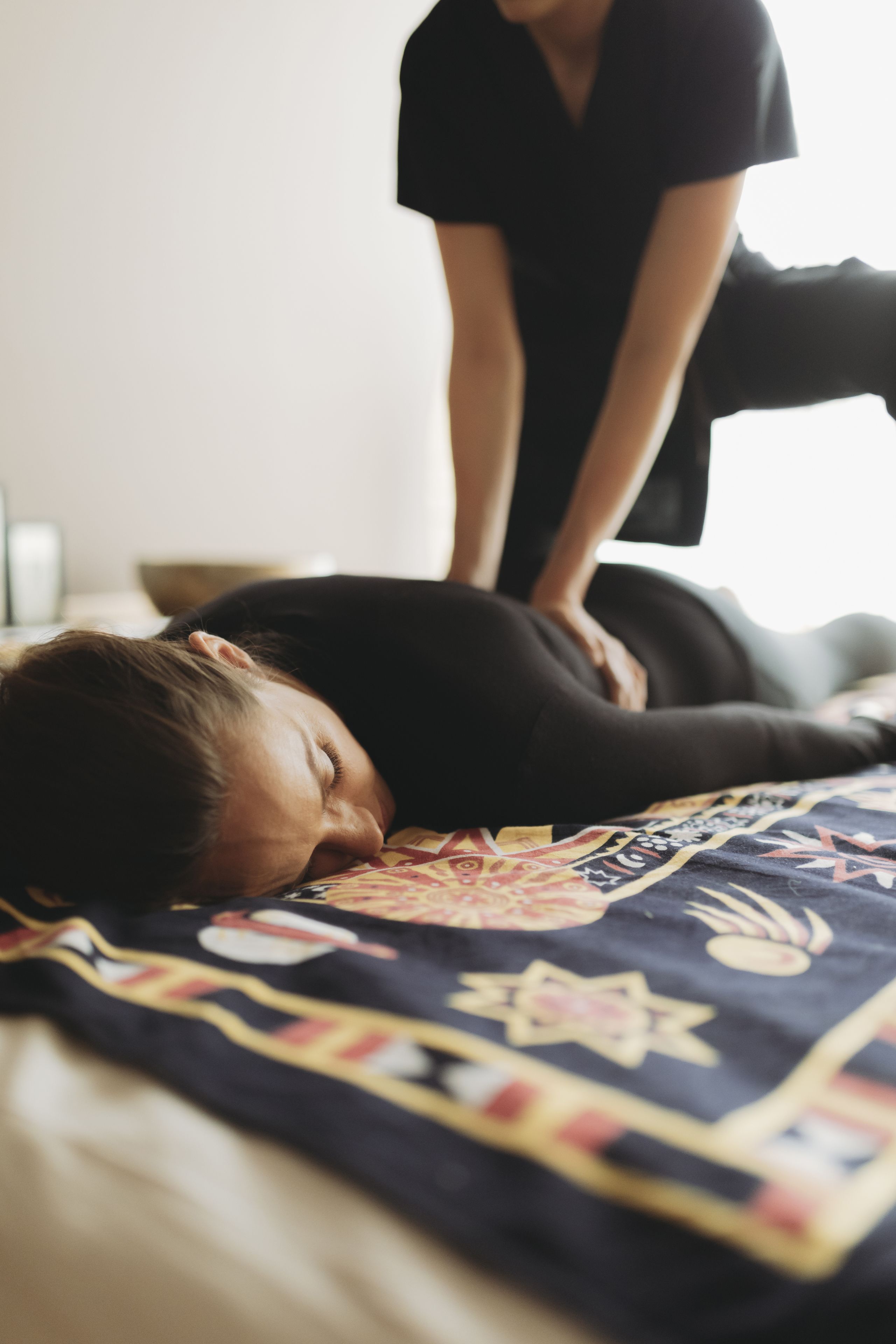 Eine Person liegt mit einer gemusterten Decke auf einem Bett, eine andere Person steht daneben, und im Hintergrund ist eine schlichte Wand zu sehen.