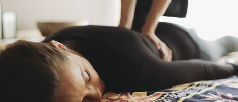 A person lying on a bed with a patterned blanket, another person standing beside them, and a plain wall in the background.