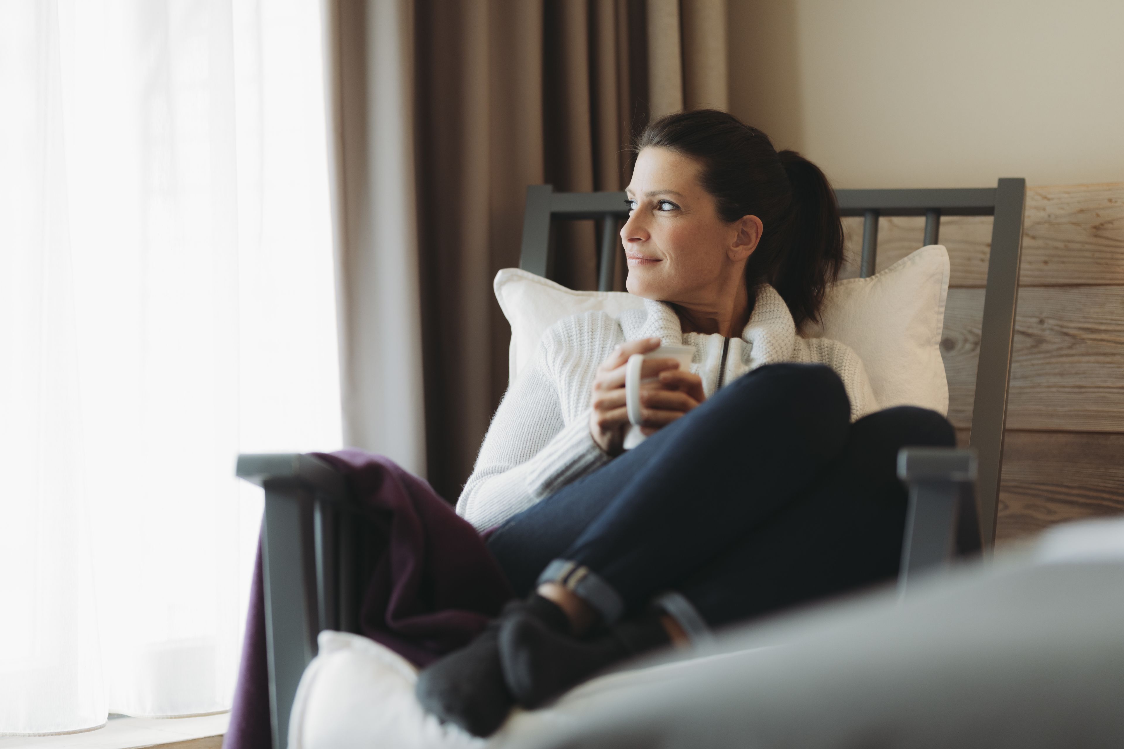A person sitting on a bed with a wooden headboard, holding a cup, in a room with a window and curtains.