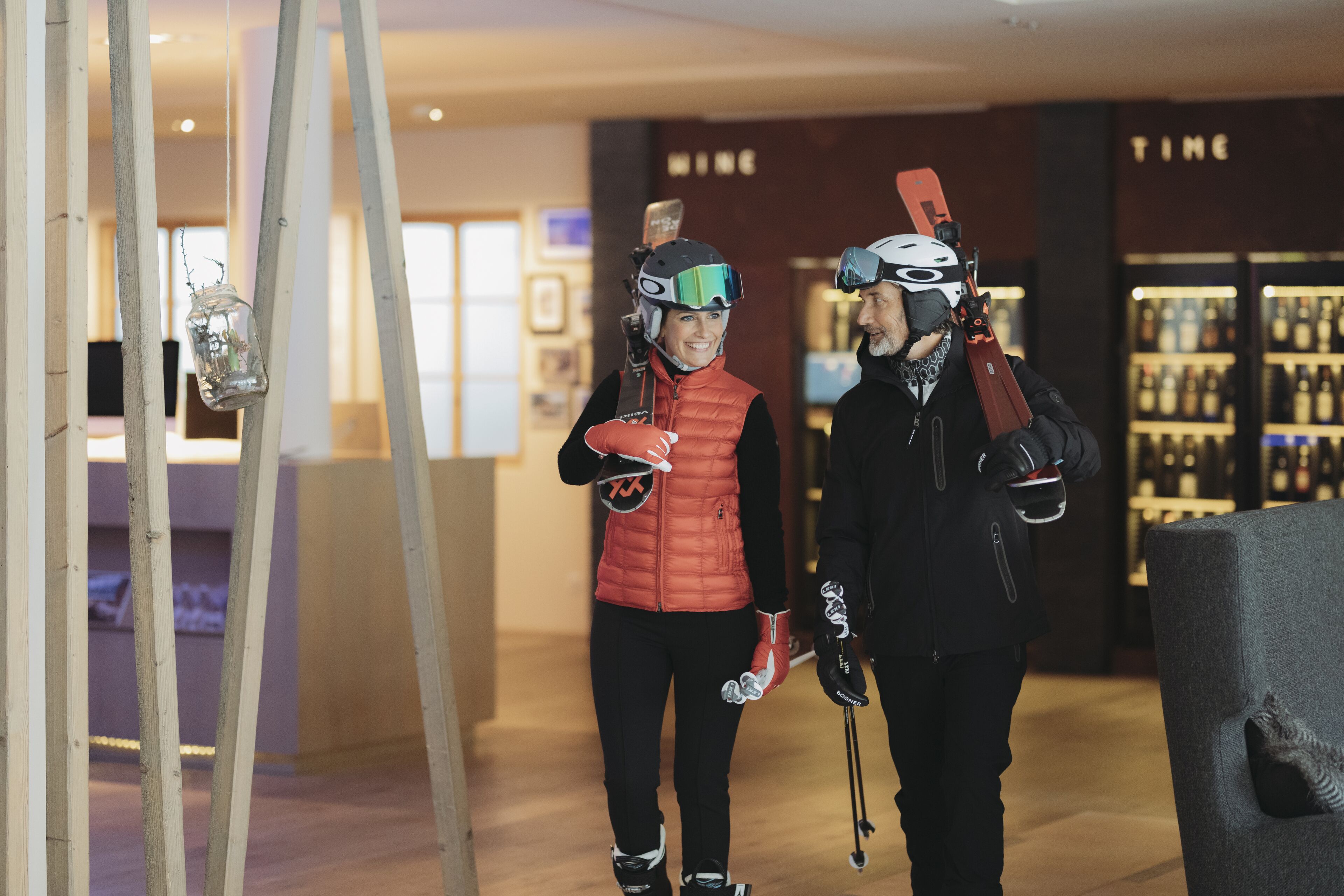 Two people in ski gear walking through a modern interior space with wooden accents and a wine display in the background.