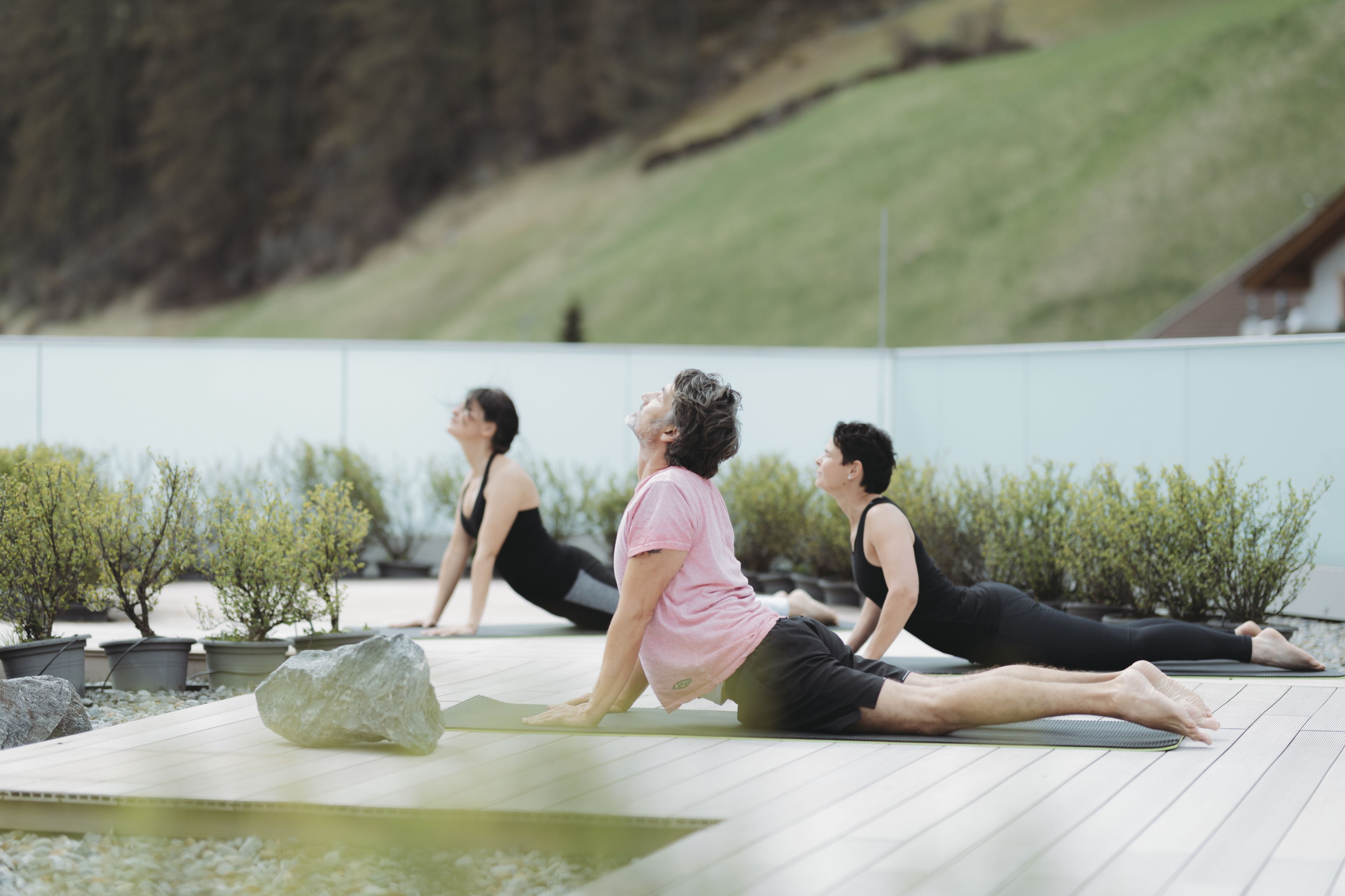 Trois personnes pratiquent le yoga sur des tapis placés à l’extérieur, dans un jardin agrémenté de plantes en pot et d’une terrasse en bois.