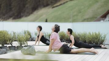 Trois personnes pratiquent le yoga sur des tapis placés à l’extérieur, dans un jardin agrémenté de plantes en pot et d’une terrasse en bois.
