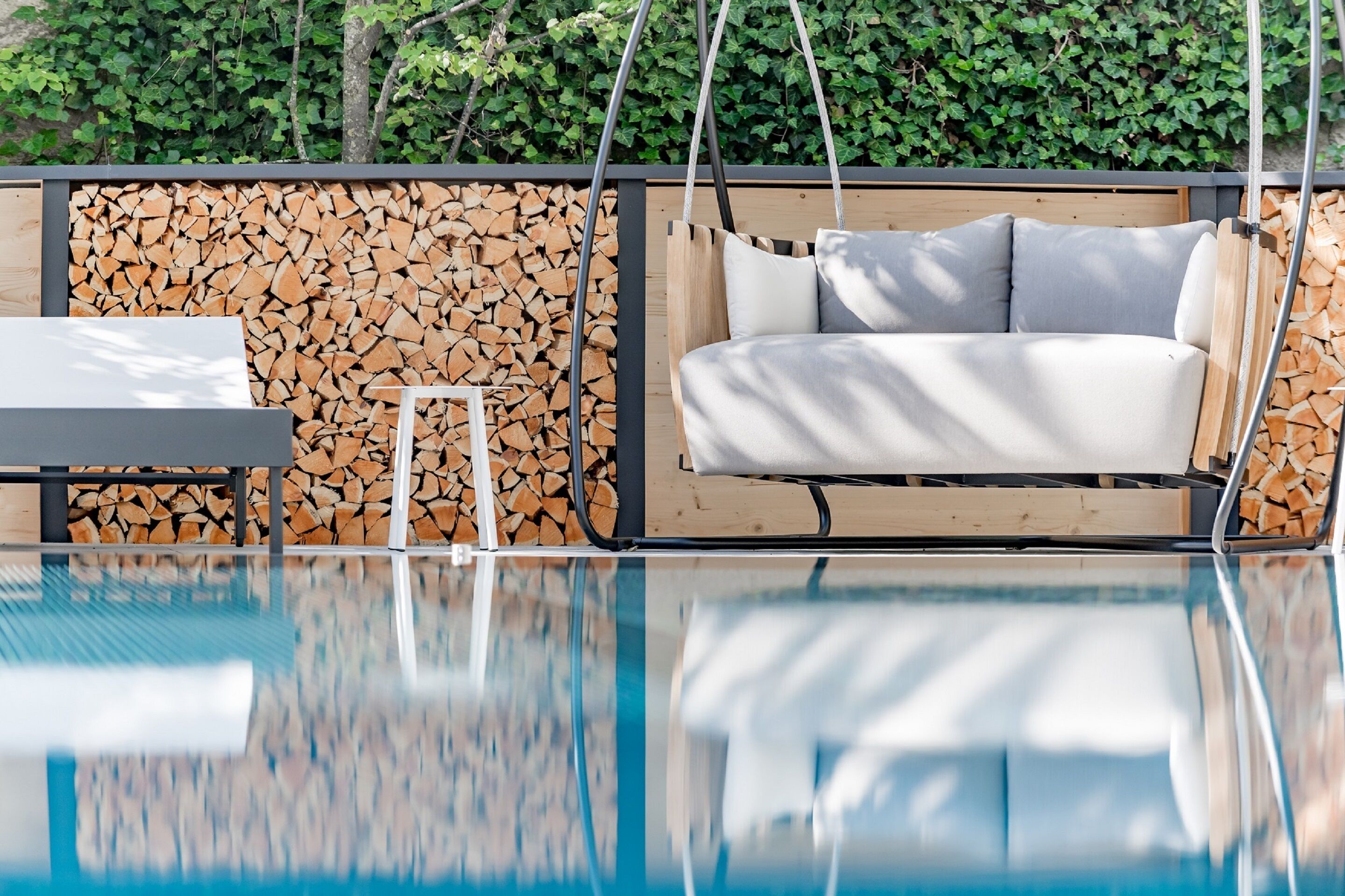 Un espace piscine extérieur moderne, agrémenté d’une chaise suspendue, d’une petite table et d’une pile de bois de chauffage adossée à un mur fait de troncs d’arbres coupés.