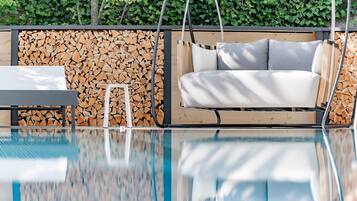 Un espace piscine extérieur moderne, agrémenté d’une chaise suspendue, d’une petite table et d’une pile de bois de chauffage adossée à un mur fait de troncs d’arbres coupés.