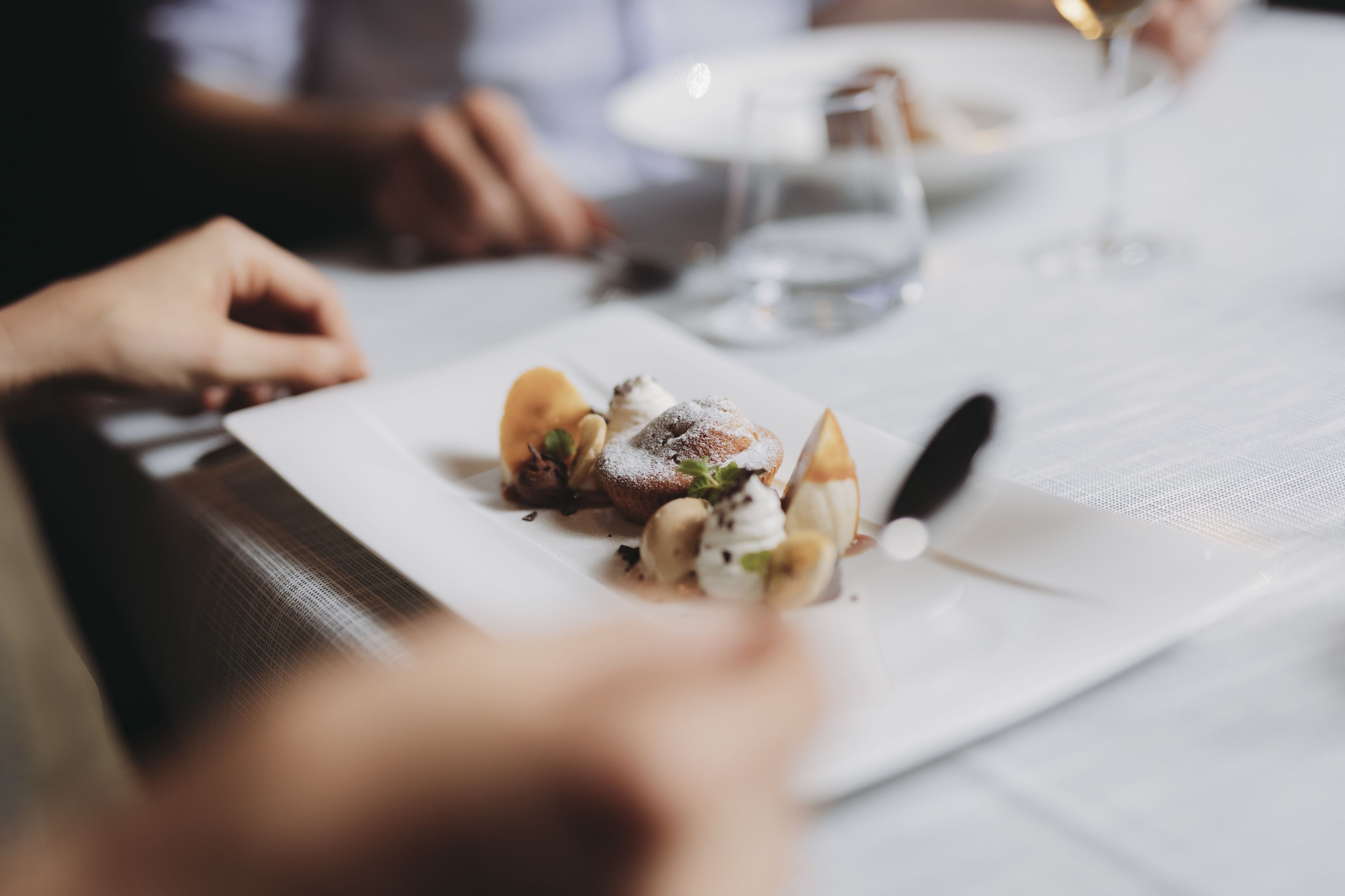 A dining table with a plate of food, a wine glass, and a person’s hands.