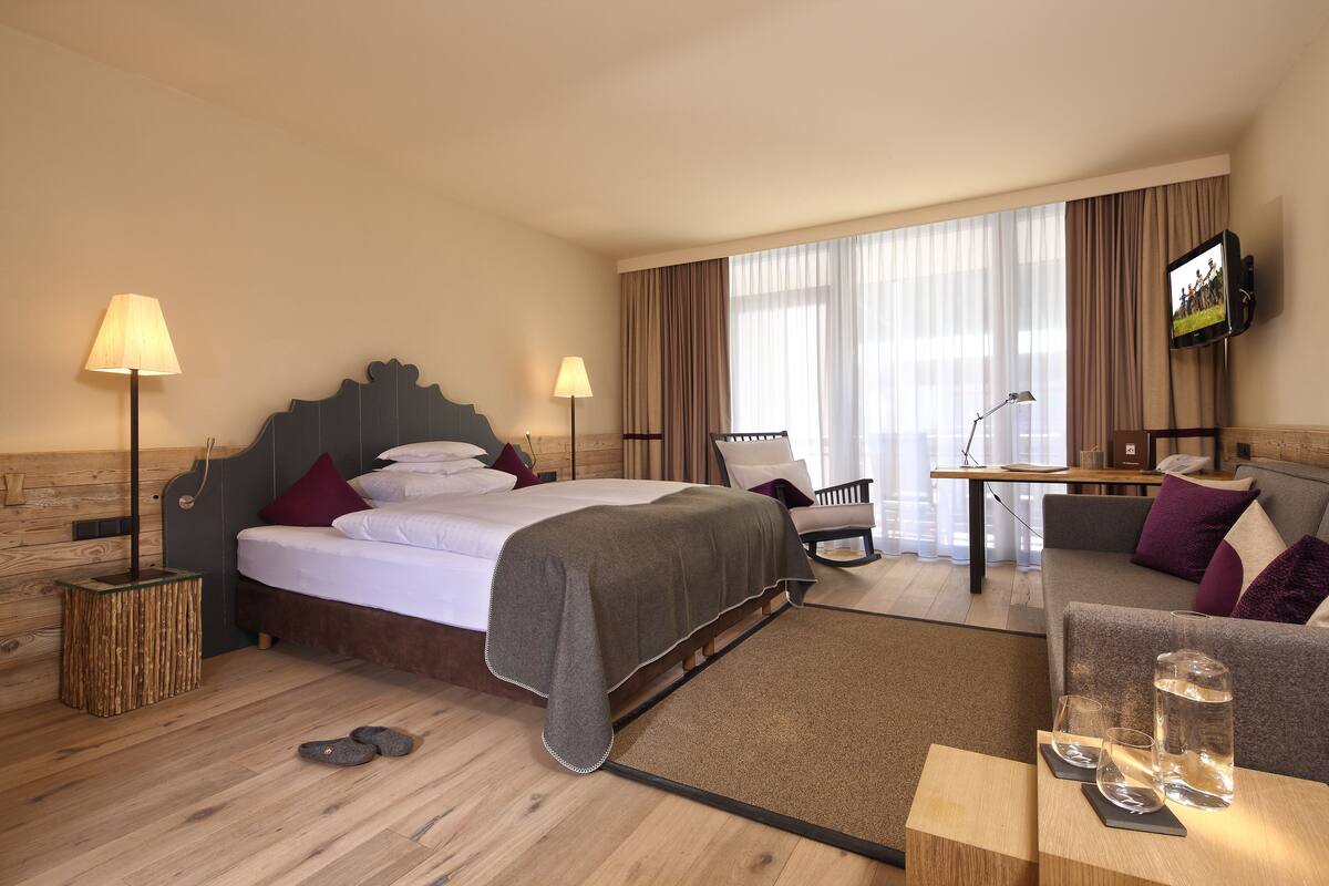 A hotel room with a bed, bedside lamps, a TV, a sofa, and a coffee table with glasses and a water bottle.