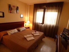 Habitación de hotel con una cama grande, una mesita de noche con lámpara, un escritorio con computadora y una ventana con cortinas.