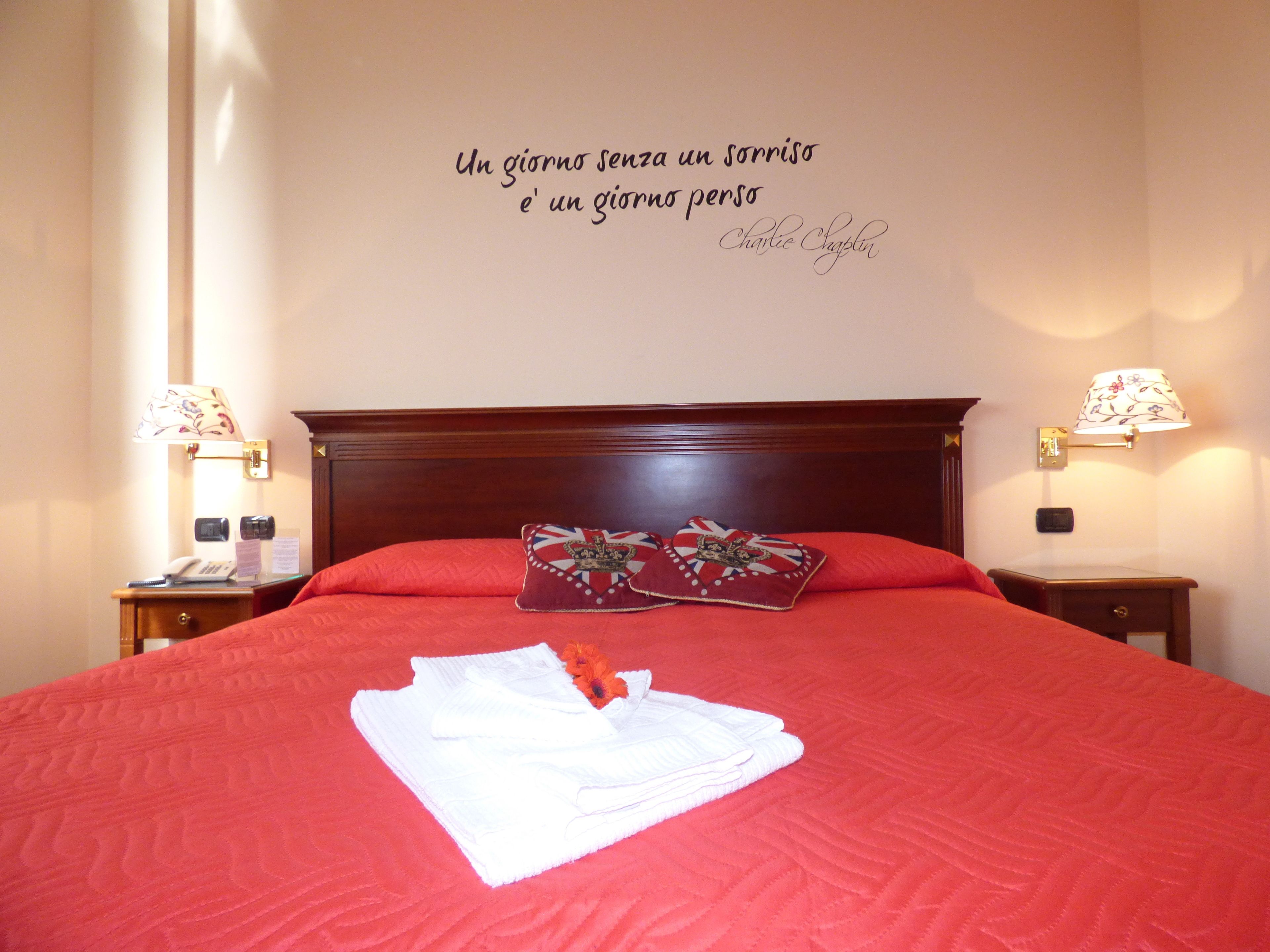 A neatly made bed with red bedding and two pillows, a bedside table with a lamp, and a wall with a motivational quote.