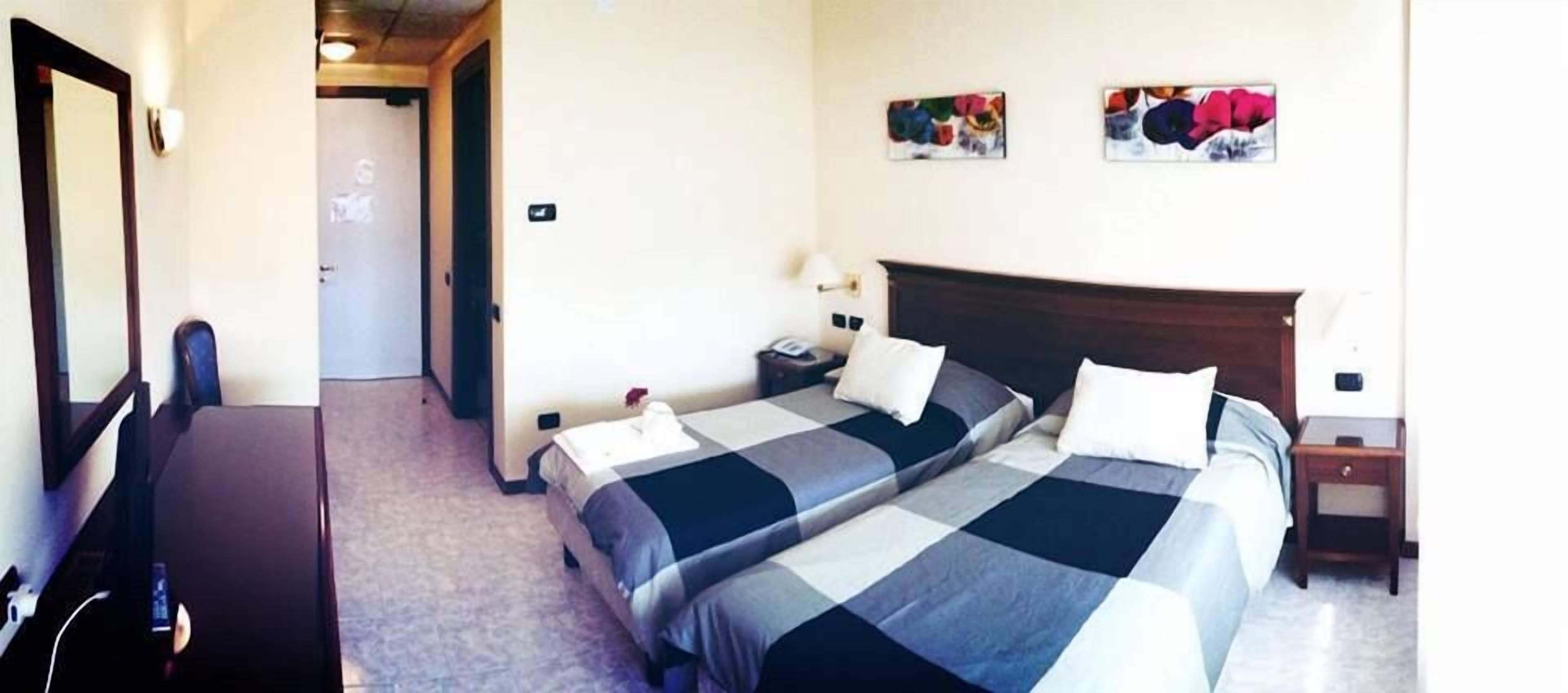 Ein Hotelzimmer mit zwei Betten, einem Schreibtisch und einem Sessel. An den Wänden hängen zwei Bilder.