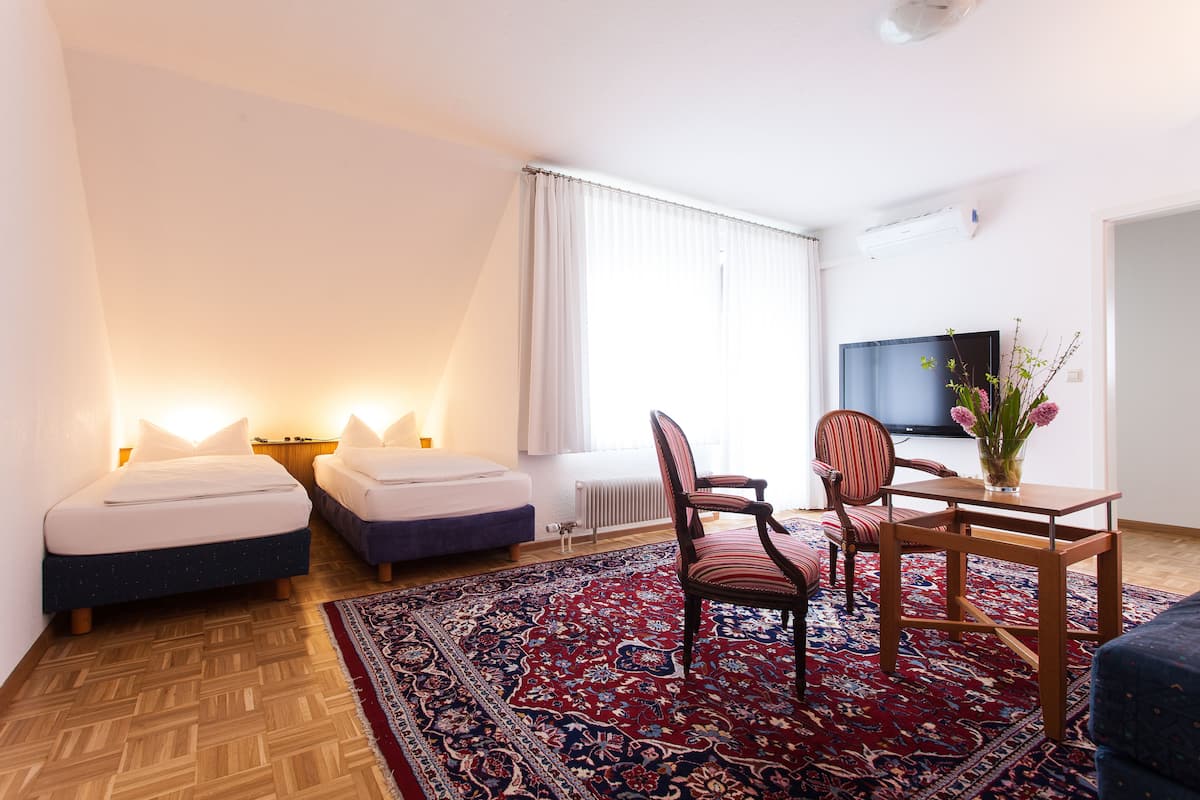 Una habitación de hotel con dos camas individuales, piso de madera, alfombra de diseño rojo y azul, una mesa de madera con una silla, un televisor y una ventana con cortinas.
