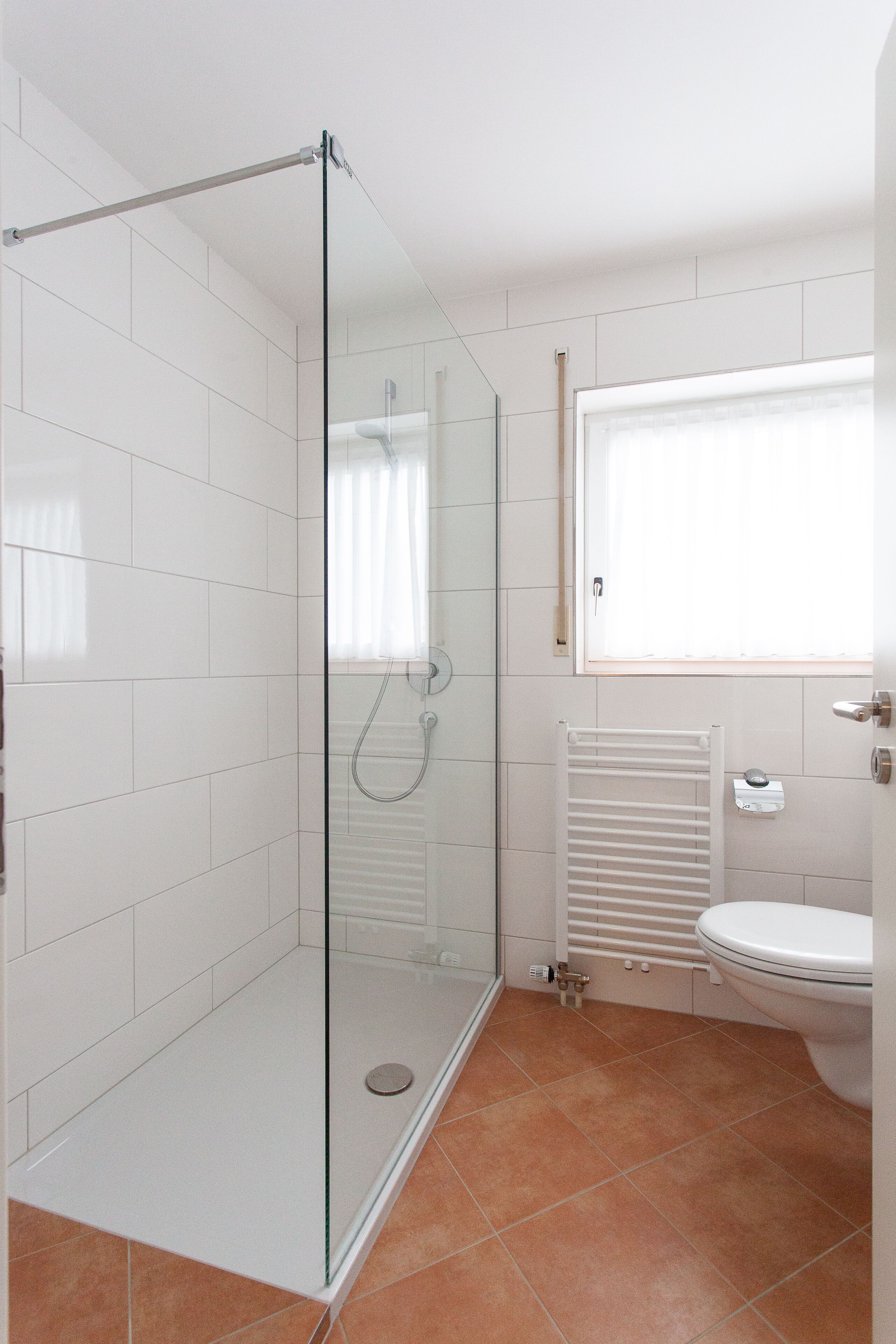 A modern bathroom with a glass-enclosed shower, white tiles, a toilet, and a window.