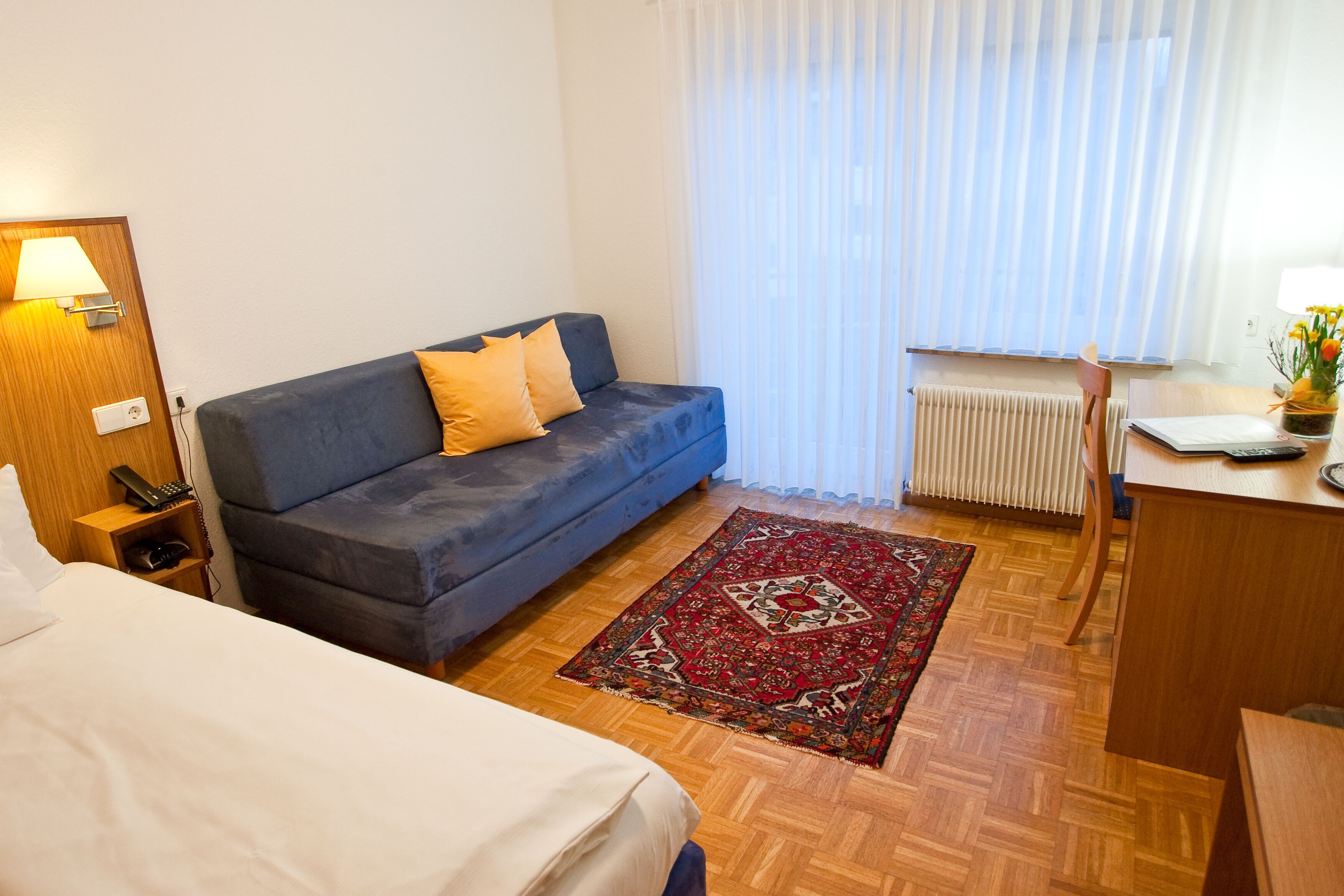 Habitación de hotel con cama, sofá azul y alfombra roja, escritorio con lámpara y ventana con cortinas.