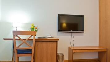 Una habitación pequeña y ordenada con un escritorio de madera, una silla, un televisor montado en la pared y un cubo de basura.