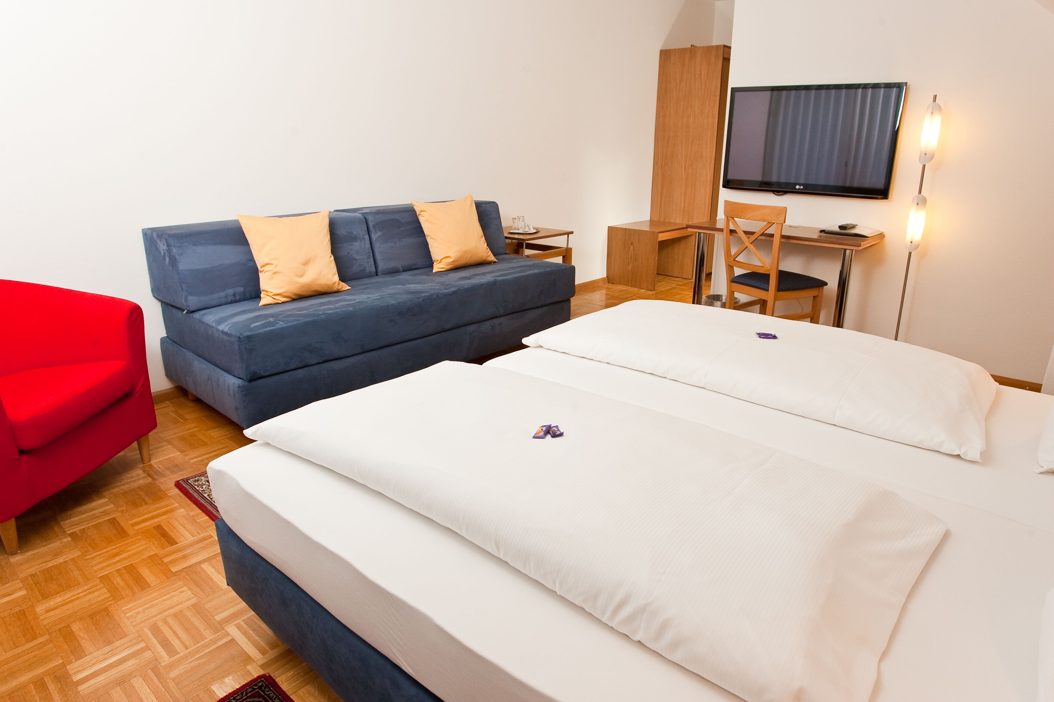 Une chambre d’hôtel avec un canapé bleu, un fauteuil rouge, un bureau avec un ordinateur, une télévision et deux lits.