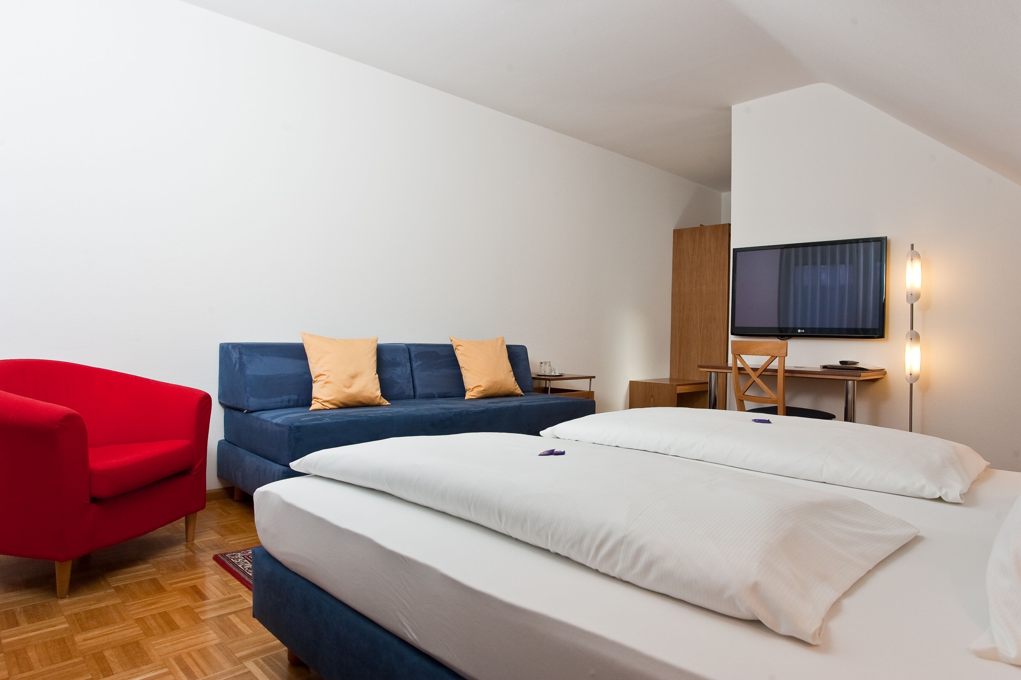 A hotel room with a bed, a red armchair, a blue sofa, a desk, a chair, and a TV mounted on the wall.
