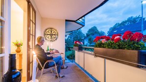 Un hombre sentado a una mesa de madera en un balcón, con una vela, una botella de vino y copas. Hay macetas con plantas y flores en la barandilla del balcón.