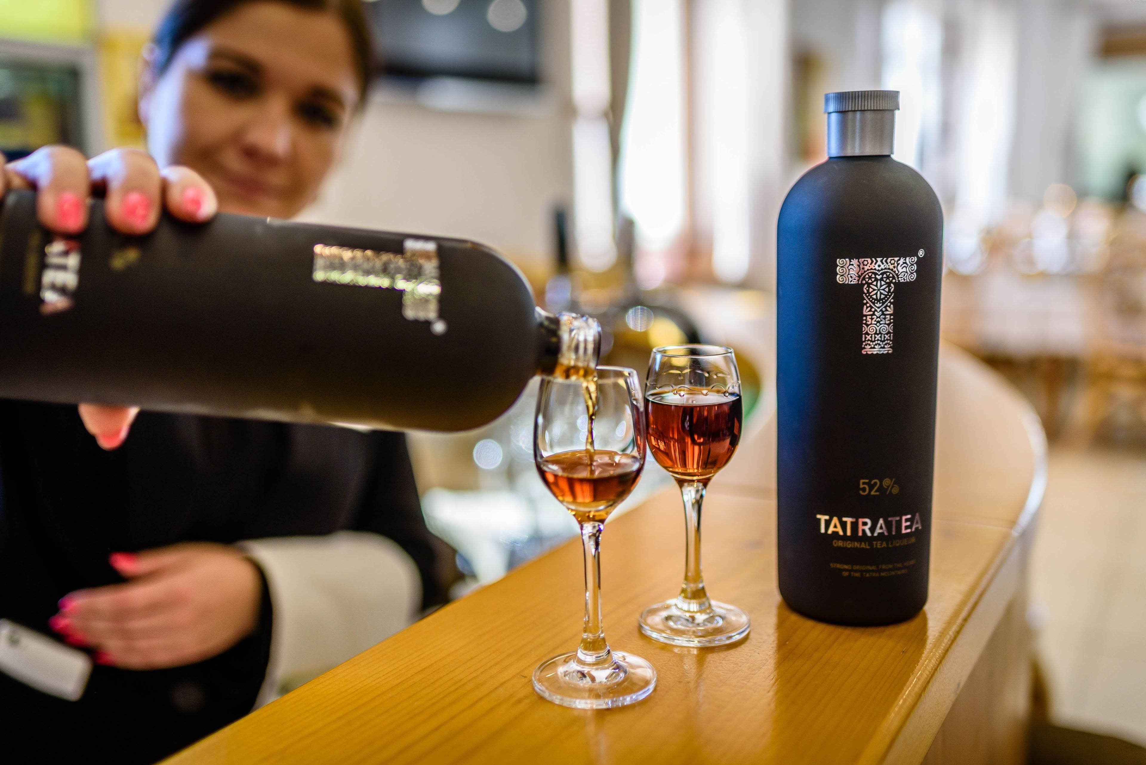 Eine Person gießt ein Getränk in zwei Gläser auf einem Holztisch, daneben eine mit 'TATRATEA' beschriftete Flasche.