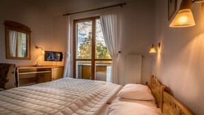 A bedroom with a large bed, a television on a wooden nightstand, a window with curtains, and a view of trees outside.