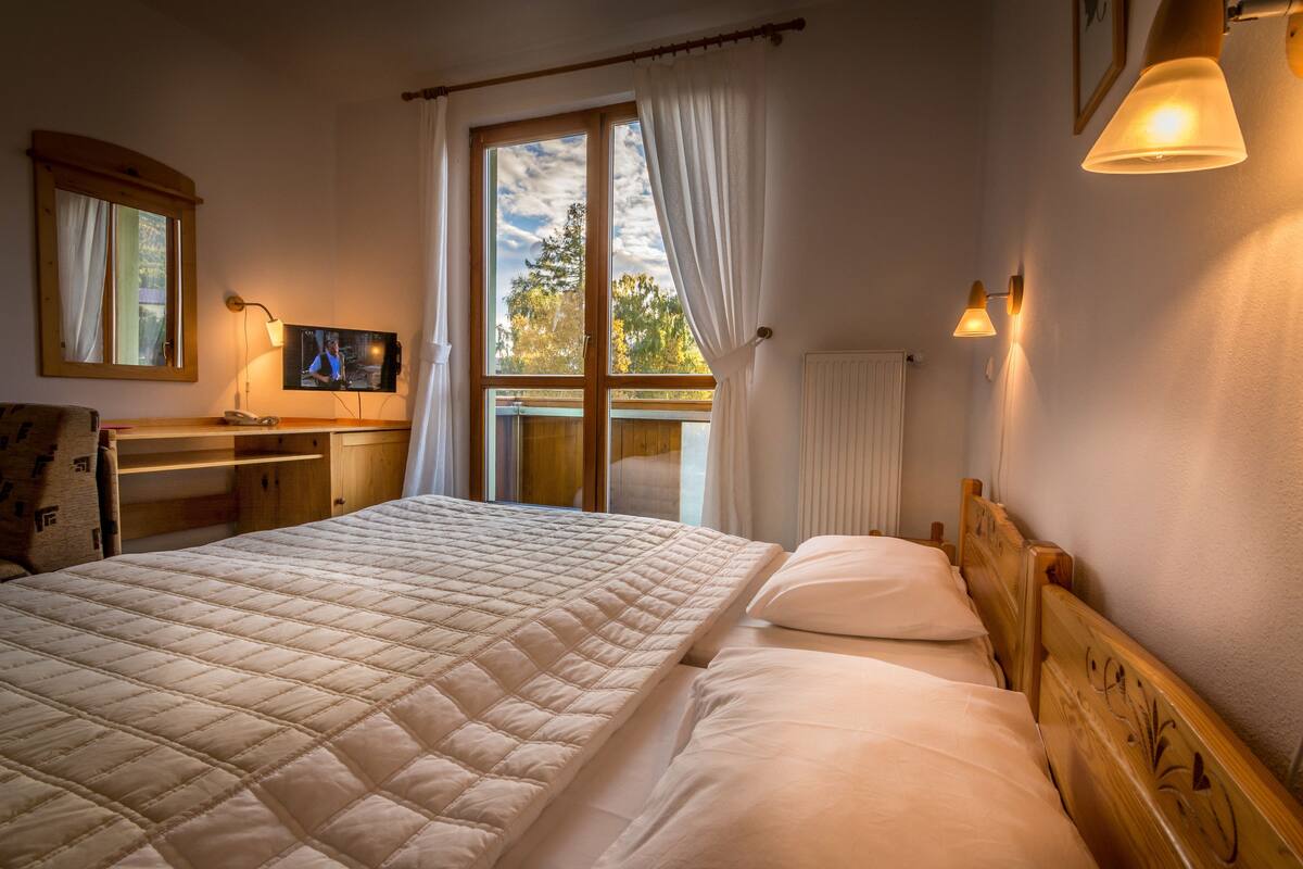 Een slaapkamer met een groot bed, een televisie op een nachtkastje van hout, een raam met gordijnen en uitzicht op bomen.
