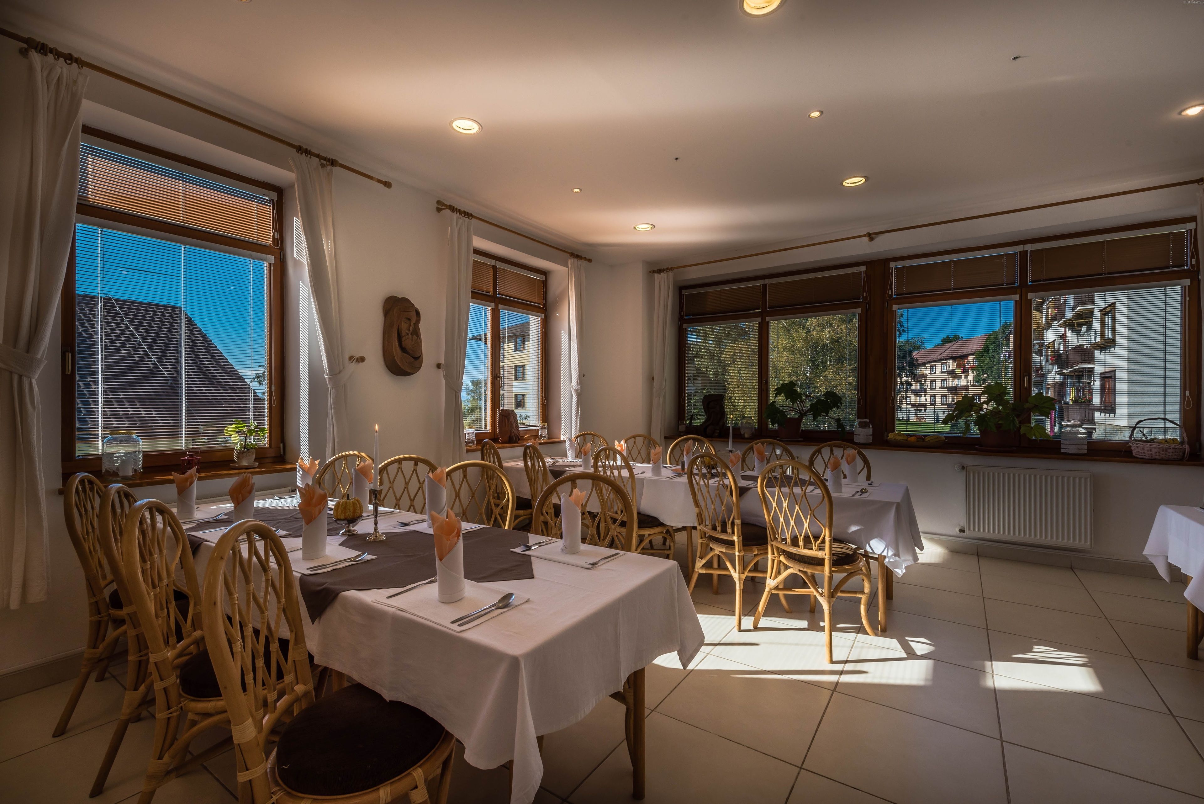 A dining area with tables set for four, wooden chairs, and large windows with blinds.