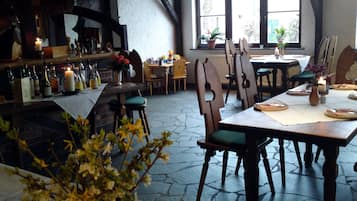 Ein Restaurant mit Steinboden, Holztischen und -stühlen. Kerzen stehen auf den Tischen, und es gibt eine Bar mit Flaschen und Gläsern.
