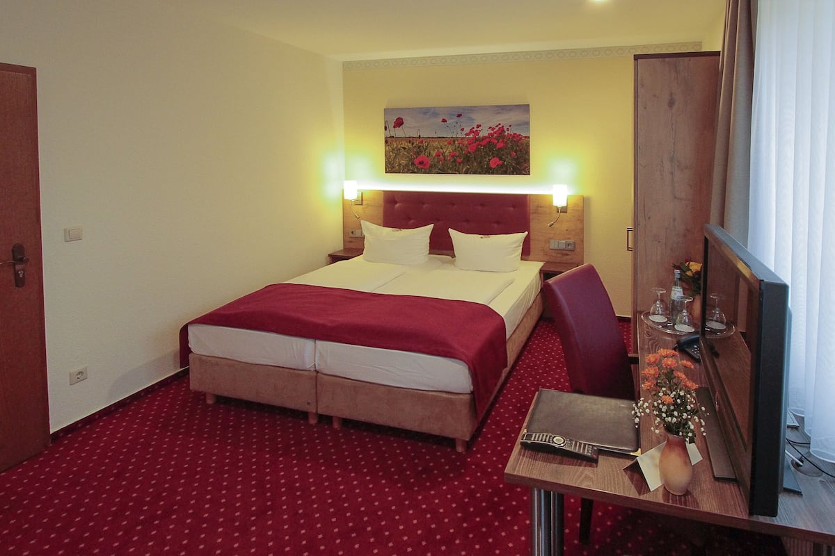 A hotel room with a double bed, a red carpet, a painting on the wall, and a side table with a vase of flowers.