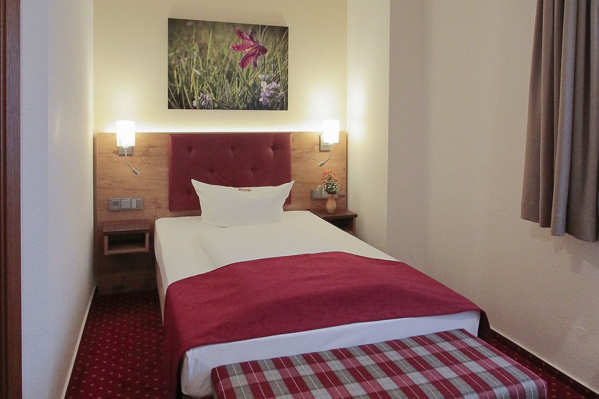 A hotel room with a bed, a bench, a nightstand, a wall-mounted lamp, and a painting.