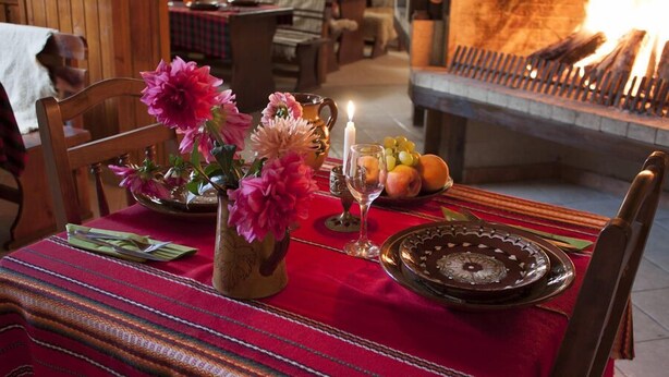 Una mesa de comedor adornada con un mantel rojo, un florero con flores, fruta y una vela.