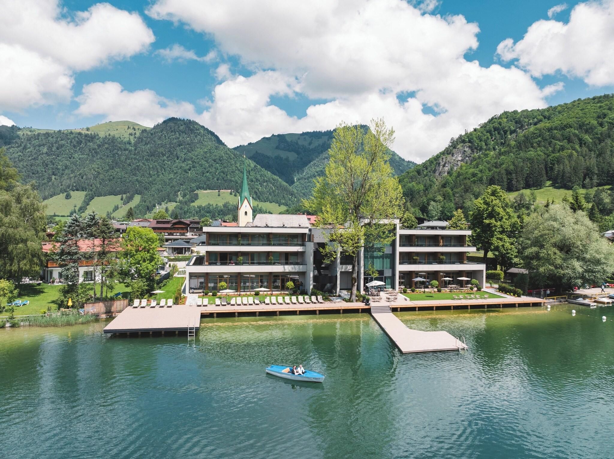 Ein Hotel am Seeufer mit Bootssteg, einer Kirche und Bergen im Hintergrund.