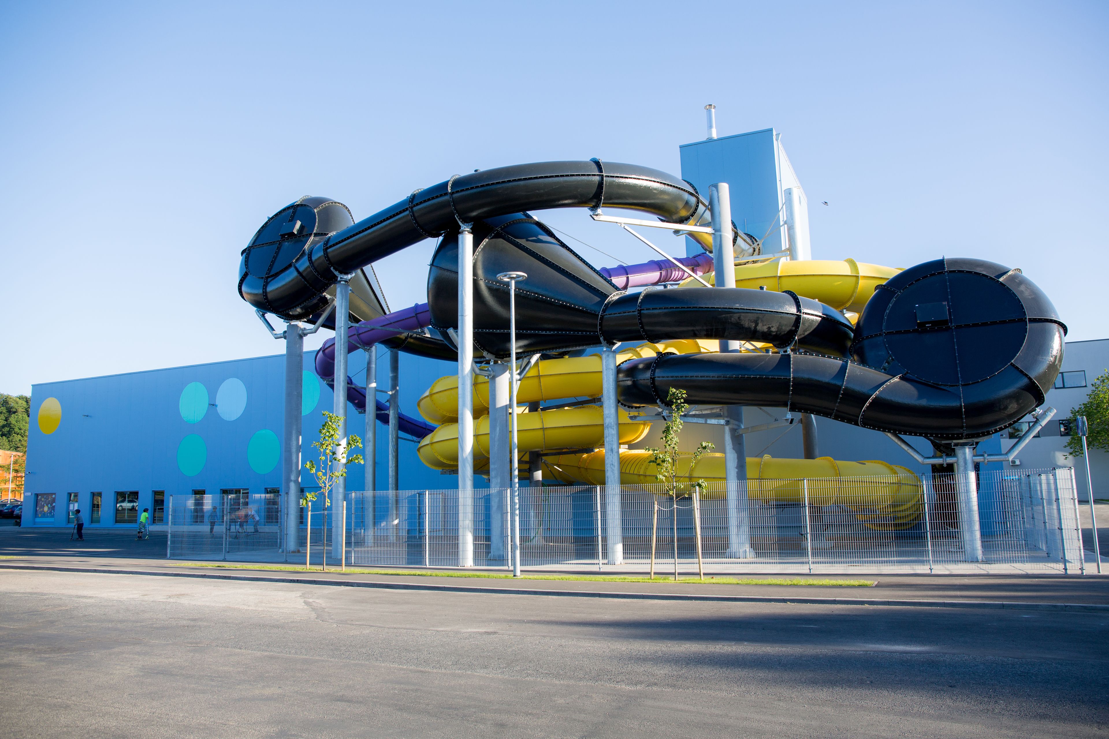 A large, colorful water slide structure with multiple loops and tubes, set against a blue building with a fence in front.
