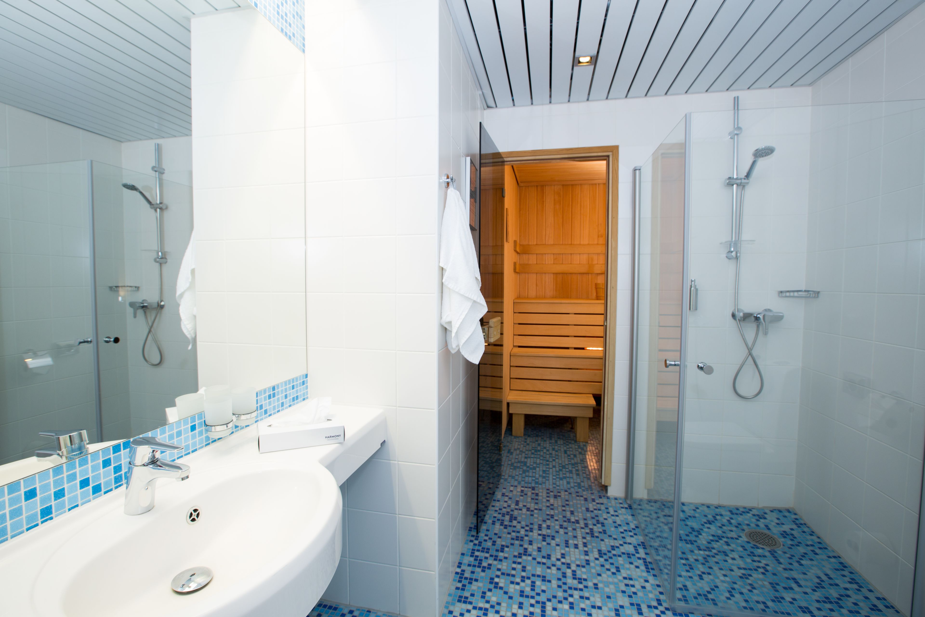 A modern bathroom with a white sink, a glass shower enclosure, and a wooden door leading to a sauna.