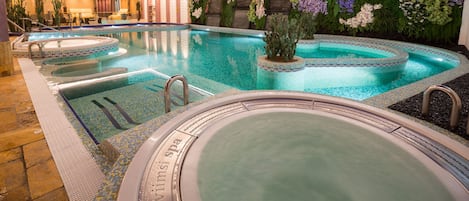 A large indoor swimming pool with multiple hot tubs, surrounded by a wall of artificial greenery and flowers.