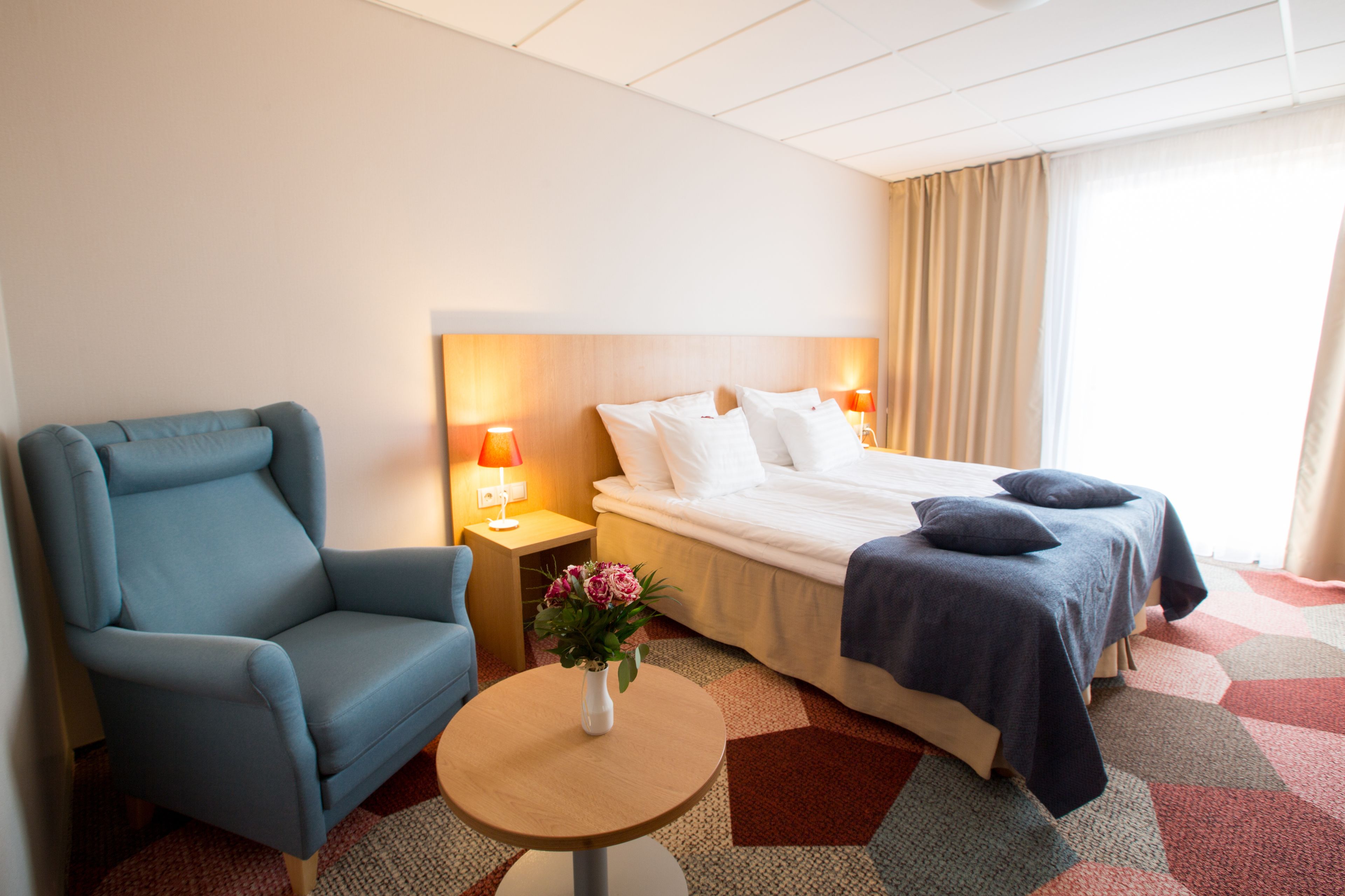 A hotel room with a bed, a blue armchair, a small round table with a vase of flowers, and a nightstand with lamps.