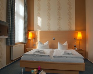 A hotel room with a large bed, two bedside lamps, a radiator, and a window with curtains.