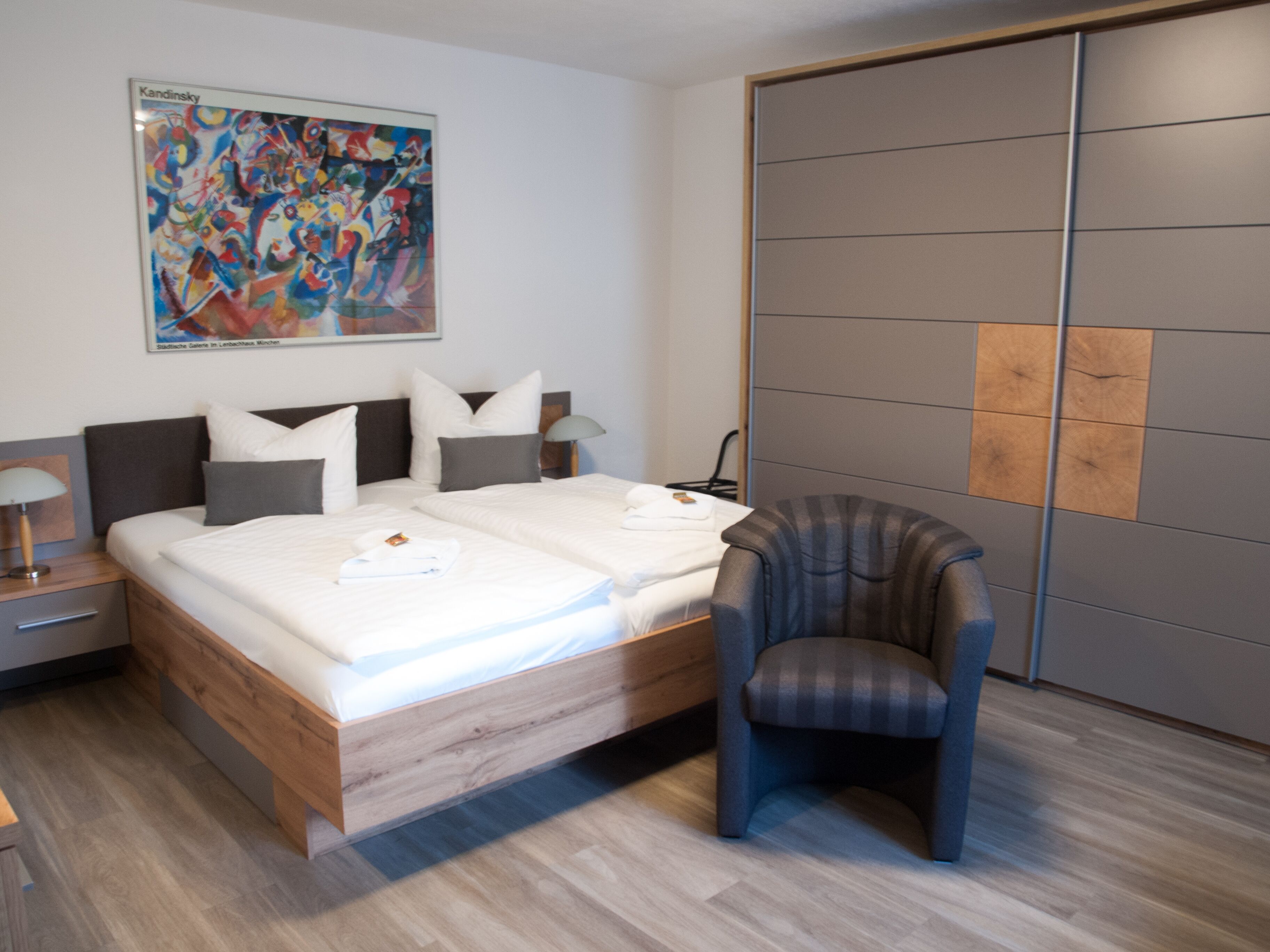 A modern bedroom with a wooden bed, a grey upholstered chair, and a large abstract painting on the wall.