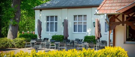 A building with a white exterior, a red-tiled roof, and a covered outdoor seating area with chairs and umbrellas.
