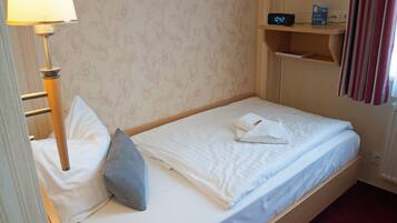 A single bed with white linens, a grey pillow, a bedside lamp, a digital clock, and a small shelf with a book.