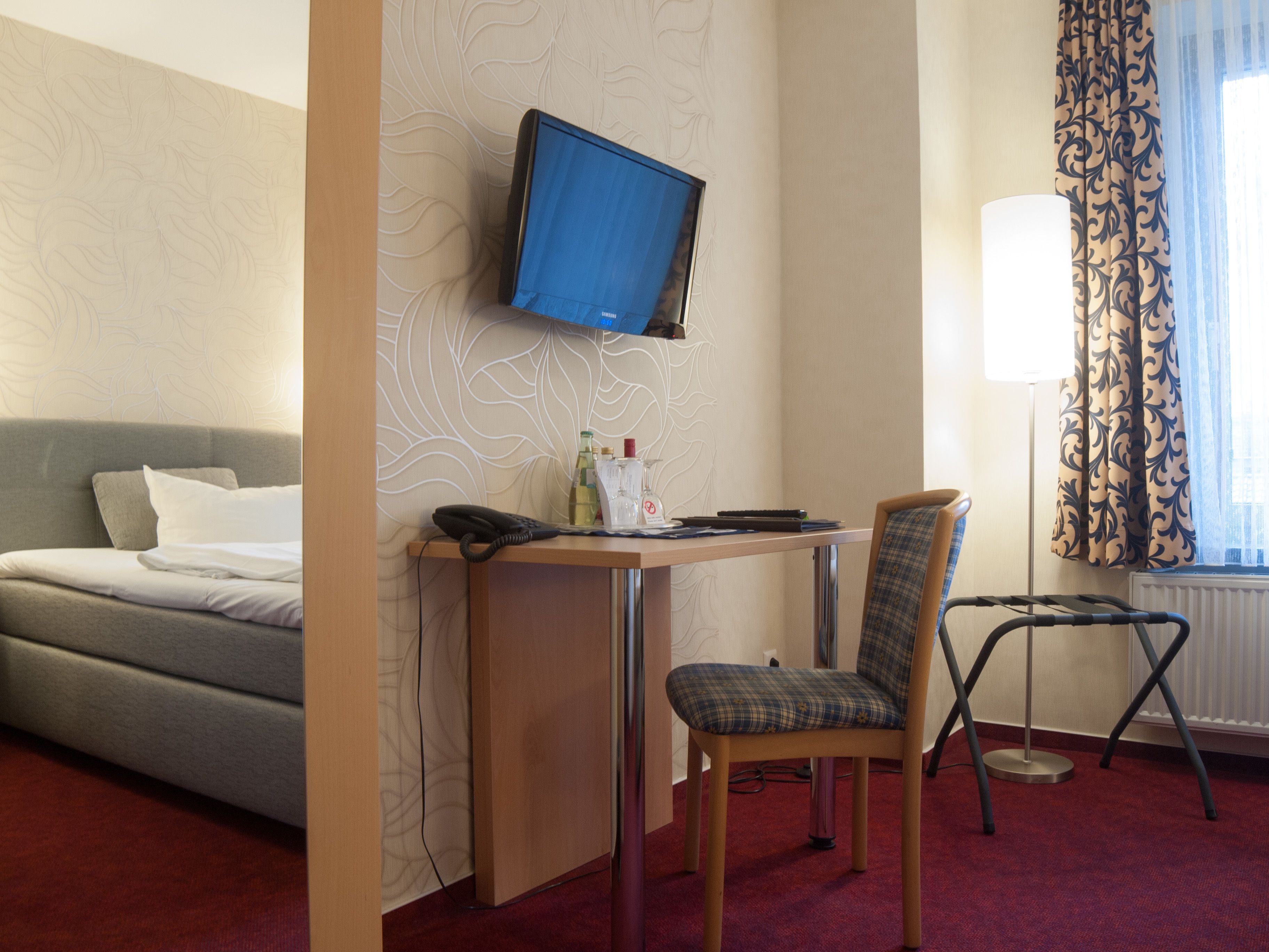 A hotel room with a bed, a desk with a chair, a television mounted on the wall, and a window with curtains.