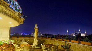 Eine Dachterrasse mit Korbmöbeln und Topfpflanzen, mit Blick auf eine Skyline, in der eine markante Moschee und Minarette dominieren.