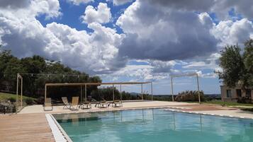 Ein Schwimmbad mit Liegestühlen und einer Pergola, umgeben von Grün und blauem Himmel.