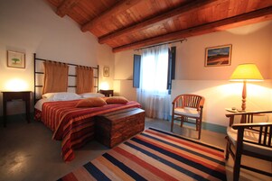 Un dormitorio acogedor con techo de madera, una cama con ropa de cama a rayas, un cofre de madera, una silla y una lámpara.
