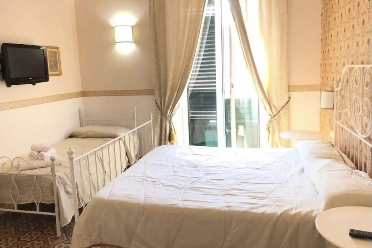 A hotel room with two single beds, a television mounted on the wall, and a window with curtains.